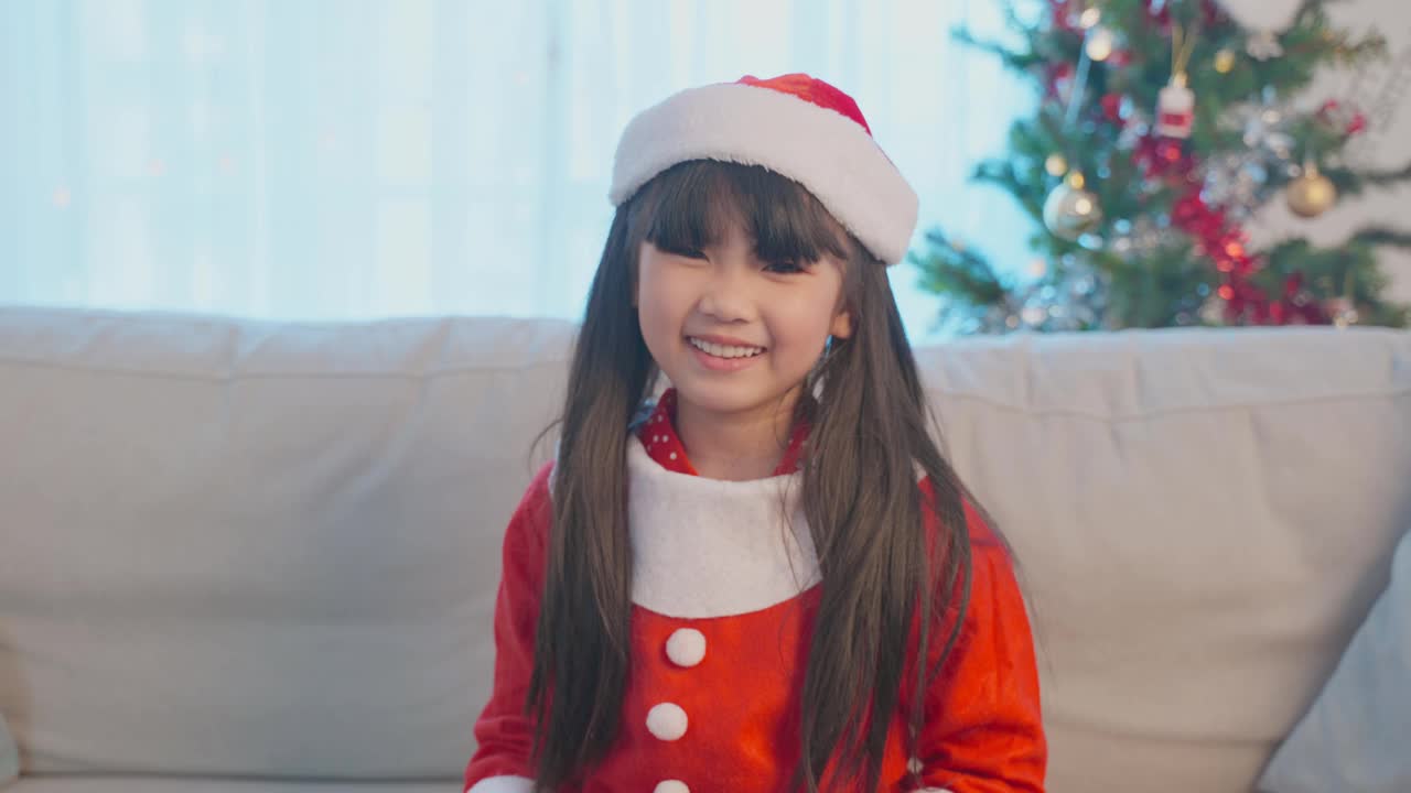 亚洲小女孩的肖像戴着圣诞帽，拿着圣诞礼物。穿着红布衣服的小可爱兴奋地庆祝节日圣诞感恩节派对，手拿礼物，看着镜头。