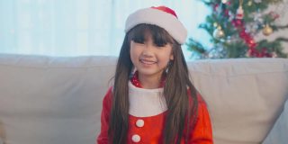 亚洲小女孩的肖像戴着圣诞帽，拿着圣诞礼物。穿着红布衣服的小可爱兴奋地庆祝节日圣诞感恩节派对，手拿礼物，看着镜头。