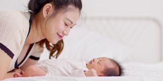 美丽迷人的亚洲妈妈亲吻宝宝的肚子甜美可爱。快乐的妈妈和宝宝一起看着宝宝，带着爱微笑。母亲和婴儿新生儿概念