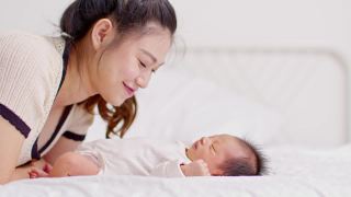 美丽迷人的亚洲妈妈亲吻宝宝的肚子甜美可爱。快乐的妈妈和宝宝一起看着宝宝，带着爱微笑。母亲和婴儿新生儿概念视频素材模板下载