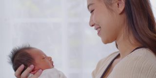 美丽迷人的亚洲妈妈抱着刚出生的宝宝，亲吻着宝宝的头，甜美可爱。快乐的妈妈和宝宝一起看着宝宝，带着爱微笑。母亲，婴儿，新生儿概念。慢动作