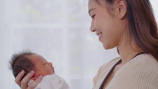 美丽迷人的亚洲妈妈抱着刚出生的宝宝，亲吻着宝宝的头，甜美可爱。快乐的妈妈和宝宝一起看着宝宝，带着爱微笑。母亲，婴儿，新生儿概念。慢动作视频素材模板下载