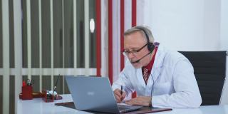 男医生与远方的病人交谈。高级医生戴着耳机，在医疗柜中的笔记本电脑上听病人进行视频聊天。