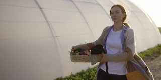 一个带着新鲜蔬菜或植物的金发女人在温室附近散步