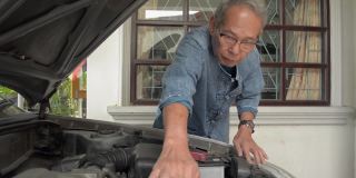一位戴着眼镜、头发灰白的亚洲老人正在家门口检查一辆旧汽车的发动机部件状况。