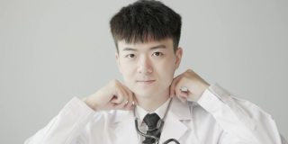 年轻的亚洲男医生，穿着白大褂，带着听诊器，对着镜头微笑，脸部特写和慢动作