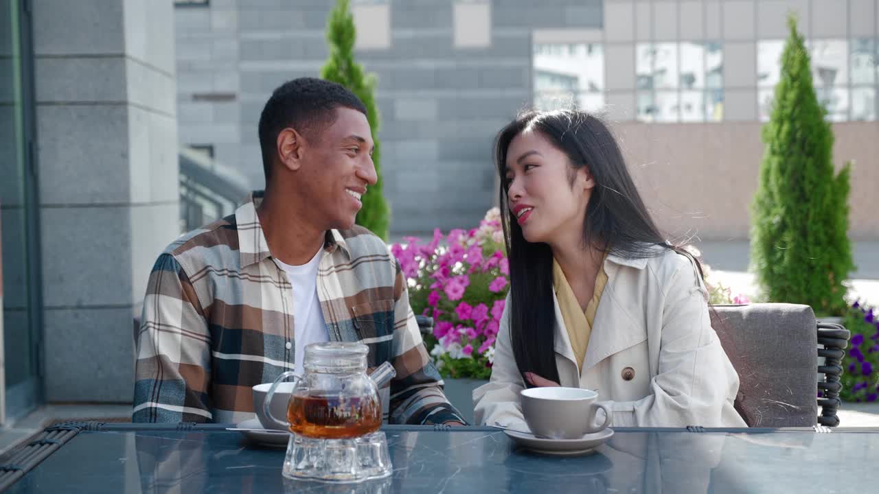迷人的年轻夫妇亚洲美丽的女人和英俊的非洲裔美国人坐在桌边与热饮料在夏天的咖啡馆，聊天，享受浪漫的约会的时刻