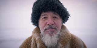 冬日里，一个留着胡子的土著老人站在户外看着镜头。老家伙头照，戴着帽子，穿着皮草大衣，表情严肃。