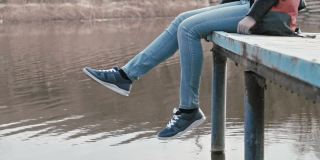 这位年轻的千禧一代女性穿着牛仔裤和运动鞋，背着背包，坐在河边的木桥边，双脚在平静的水面上摆动。重建的概念。缓慢的运动。
