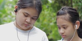 两名泰国少女在公园里戴着耳机，用智能手机观看社交视频。在兄弟姐妹家庭中团结在一起。农村青年的现代生活。