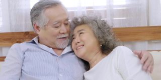 亚洲健康的爷爷坐在客厅的沙发上互相拥抱表示爱，欢快的老夫妇在房子里微笑