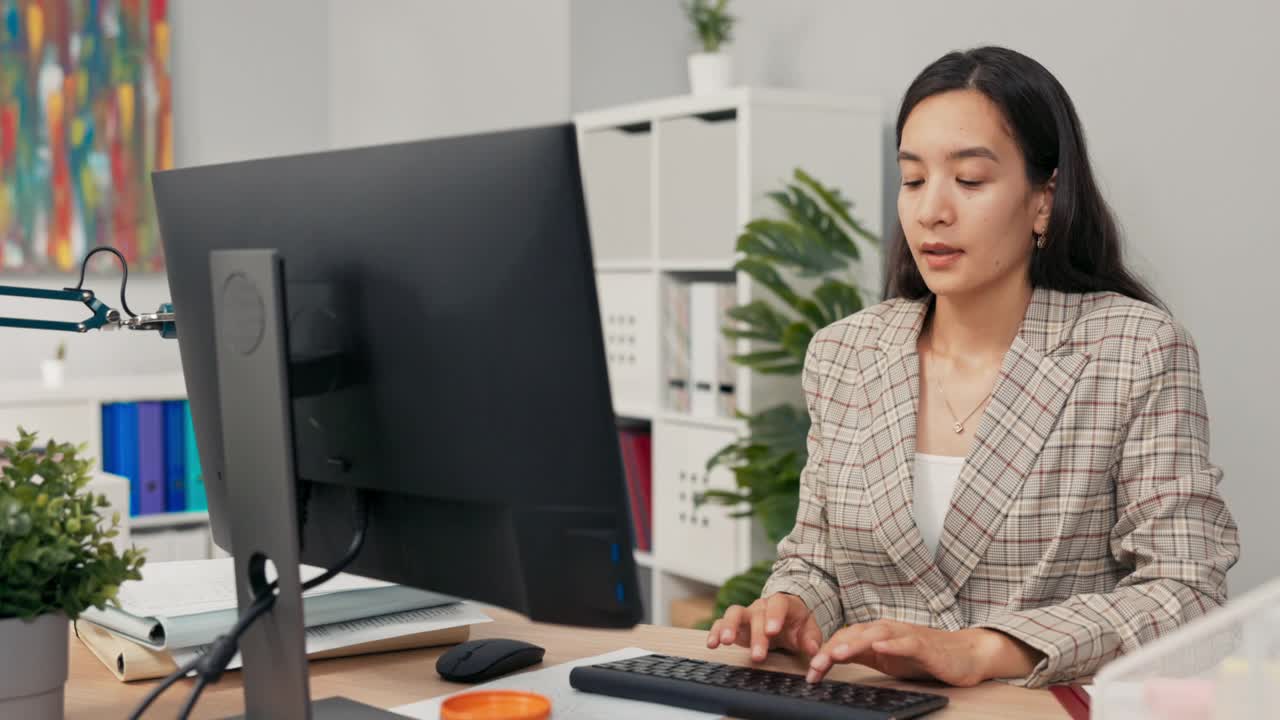 一名棕色长发穿着格子夹克的女子坐在办公室电脑屏幕前，通过网络摄像头接收电话，与同事视频聊天，展示她需要修改的文件