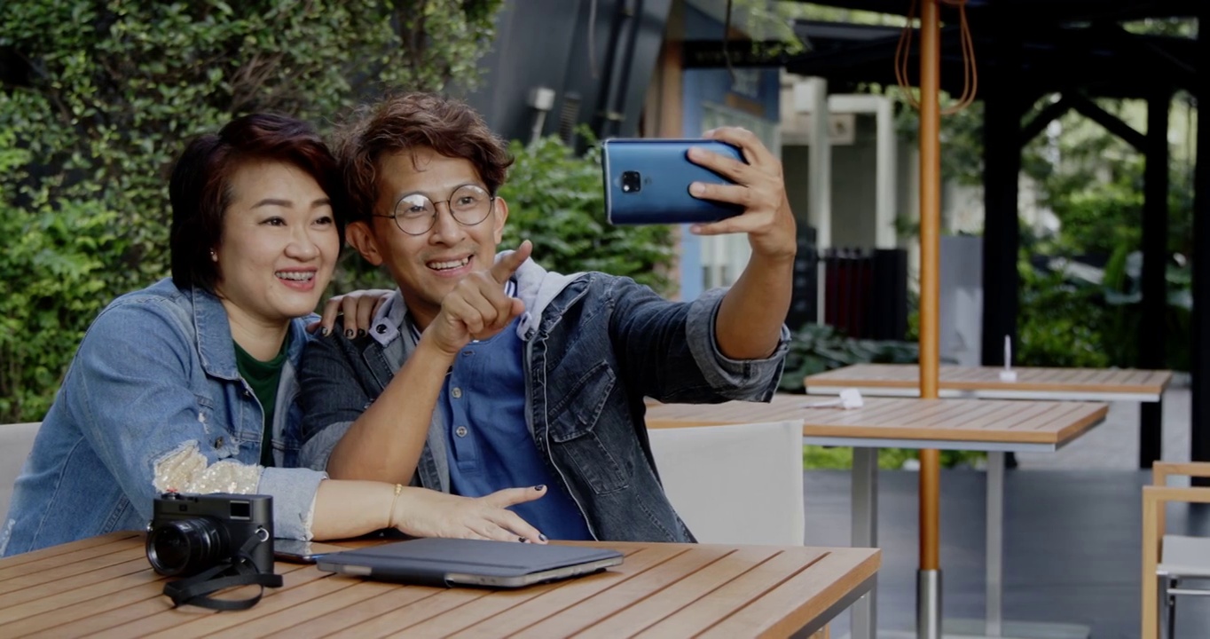 亚洲家庭情侣在夏日花园度假时用智能手机自拍。带着快乐的微笑坐在公园里视频聊天。休闲活动开放后的城市独立生活