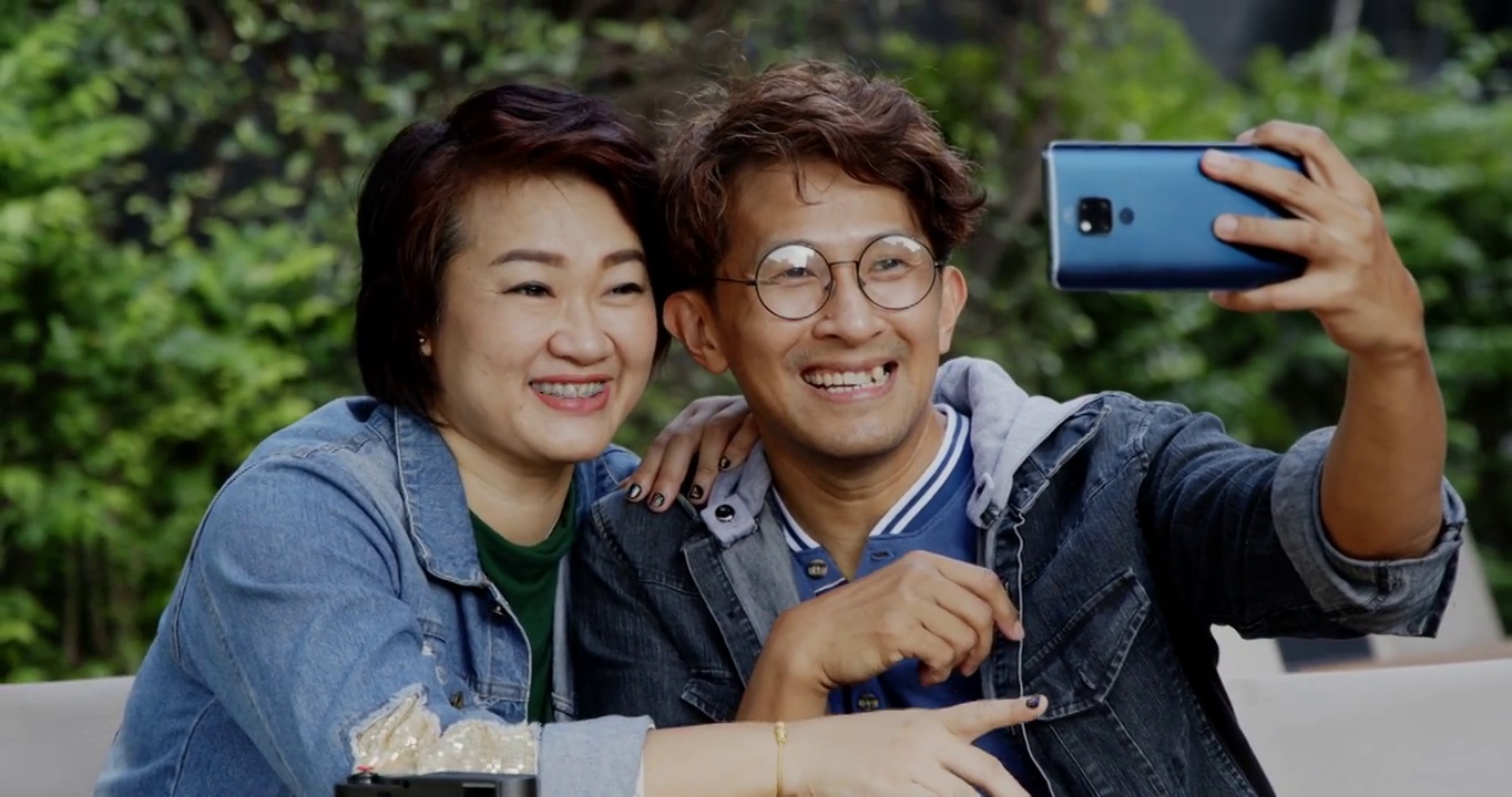 亚洲家庭情侣在夏日花园度假时用智能手机自拍。带着快乐的微笑坐在公园里视频聊天。休闲活动开放后的城市独立生活