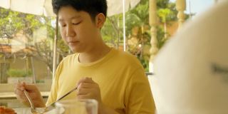 快乐的亚洲男孩在酒店的窗口附近吃早餐在他的假期旅行与家人，生活方式的概念。