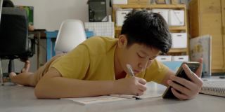 亚洲男孩躺在地板上做作业，同时在家学习在线课程，社交距离的生活方式概念。