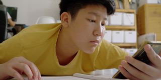 亚洲男孩躺在地板上做作业，同时在家学习在线课程，社交距离的生活方式概念。