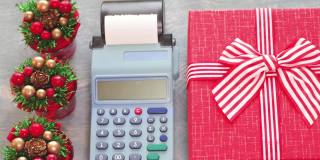 圣诞节销售。收银员的手在收银机上数着美元现金，旁边是一个红色的礼品盒和小圣诞树，俯视图。购物新年和圣诞节礼物特写，全高清