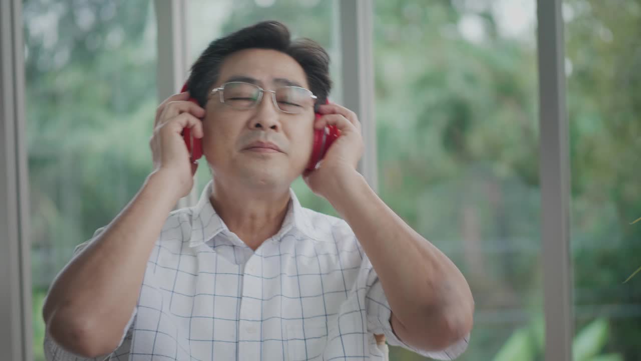 年迈的亚洲男人在家里的客厅里享受着业余爱好，戴着耳机听着音乐，脸上挂着快乐的笑容