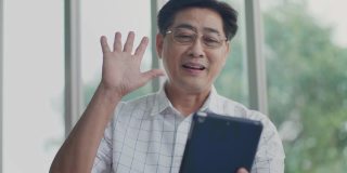 一个亚洲老人坐在房子里。享受使用平板电脑通过应用程序与家庭视频通话。概念通信技术