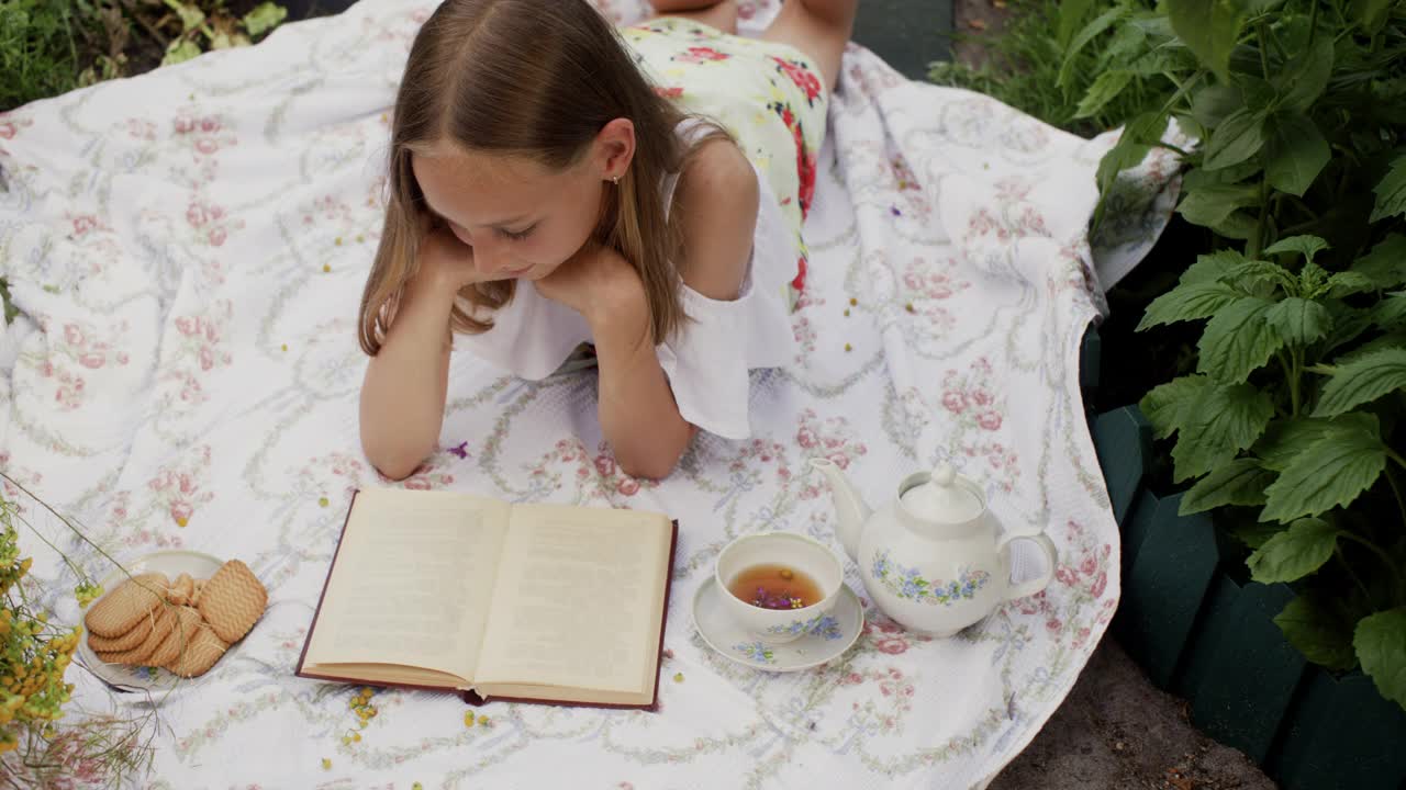 年轻女子躺在夏天的草地上看书，地毯上放着饼干和茶。浪漫的女人拿着书、茶和饼干在夏日的花丛中放松。