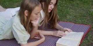十几岁的女孩在暑假躺在绿色的草坪上看书。两个女朋友在夏日公园的草地上悠闲地看书。