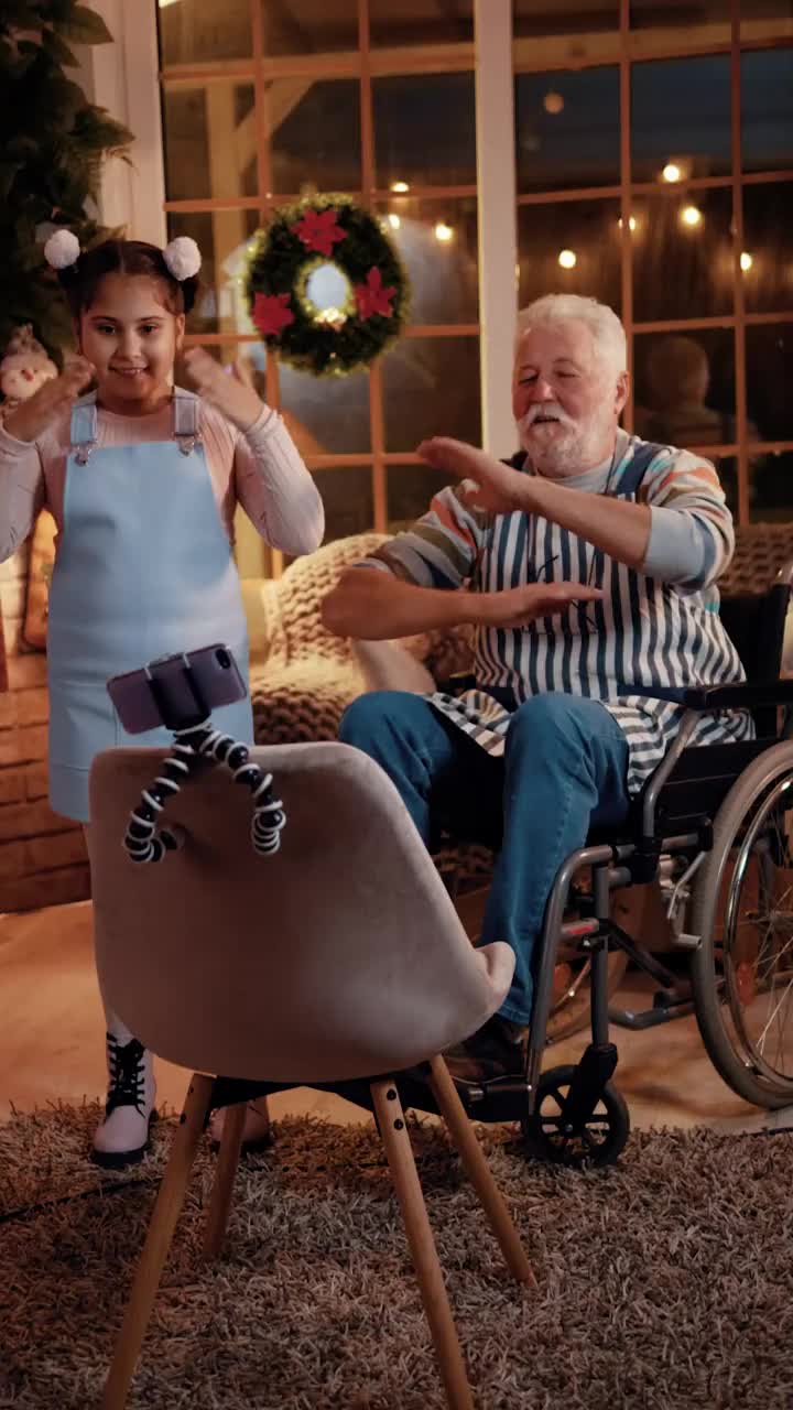 一个残疾的爷爷和一个小女孩在家里为社交网络跳舞。家人在家里用智能手机在社交媒体上跳舞和直播。垂直