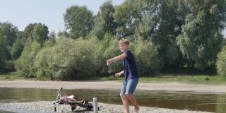 一个可爱的白人少年赤脚站在河边往水里扔石头。自然的休闲活动。缓慢的运动。