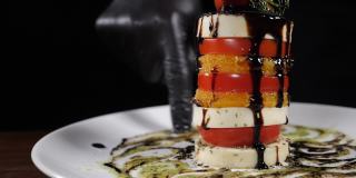 慢动作食物视频概念。白盘子里的沙拉加香醋。切成片的马苏里拉奶酪，辣椒酱，番茄，一个接一个。厨师用手指在盘子上涂抹酱汁。全高清
