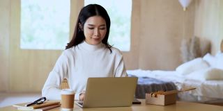美丽微笑的亚洲创意女性穿着毛衣，一边在家里用笔记本电脑工作一边讲手机。年轻的亚洲自由职业者妇女在家工作。小企业主。
