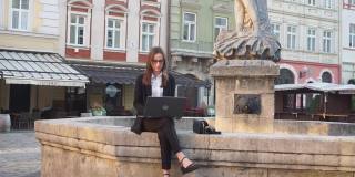 一个小女孩坐在外面用笔记本电脑。美丽的年轻女子坐在城市中心的喷泉边，带着笔记本电脑在网上聊天