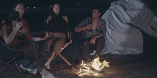 一群亚洲年轻人在野外露营。年轻人晚上在营火旁一起弹吉他唱歌
