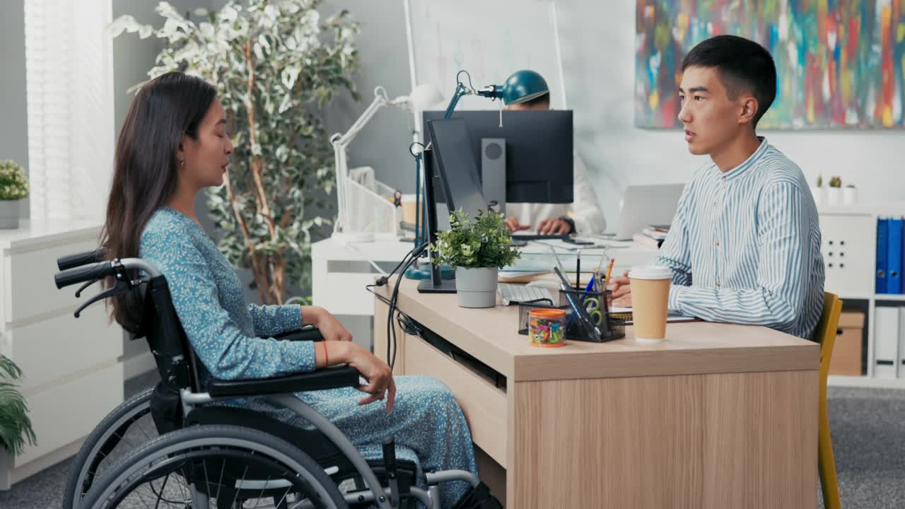 与坐轮椅的残疾妇女求职面试，亚裔美女严厉的男老板向穿着衣服的女孩提问，他们坐在公司办公室办公桌两侧的小伙子仔细听着回答