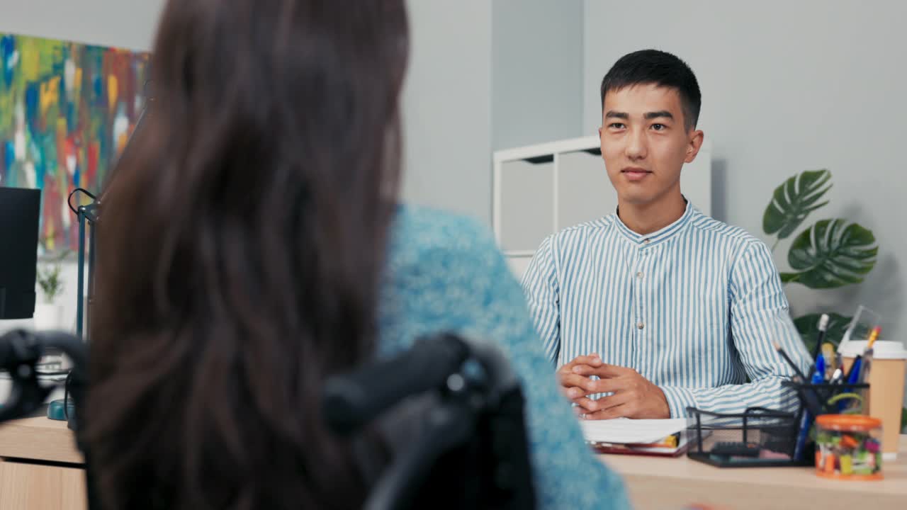 衬衫男手拿纸坐在桌子后面，老板问隐形轮椅上的女人问题，雇残疾女孩当秘书，阅读合同，在电脑里检查女性资料