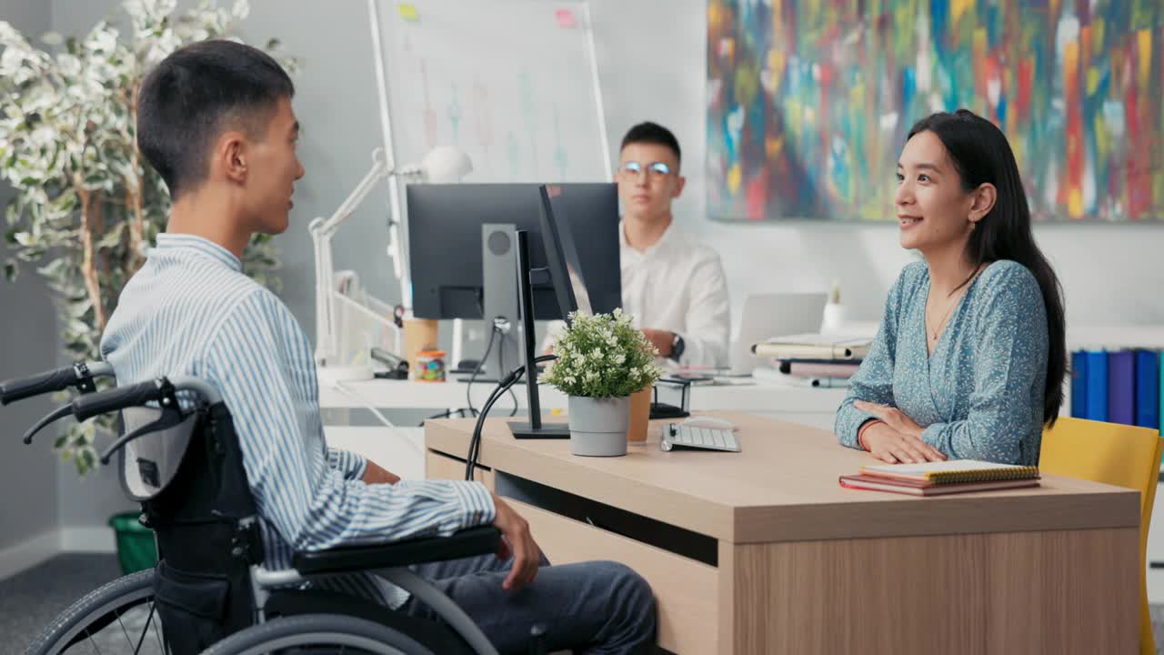一个坐着轮椅的残疾人面试，一个亚裔韩国美女向穿着衬衫的男孩提问，她听着他们坐在公司办公室桌子两边的答案