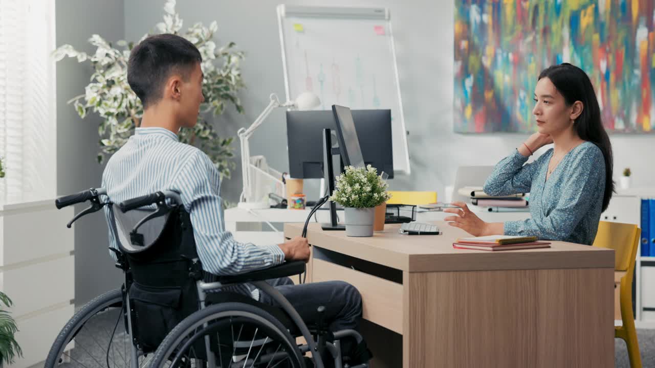 一个男人坐在轮椅上坐在办公室的桌子前，他对面是一个穿着裙子的漂亮女人，他们签署了一份合作雇佣协议，感谢对方的面试，祝贺