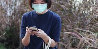 亚洲女性戴着防护面具在户外花园玩智能手机放松