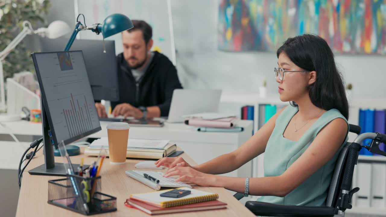 工作日在办公室，公司，公司，同事们都在电脑前工作，起草文件，一个戴着眼镜的亚洲美女坐在轮椅上，她是残疾人，事故后