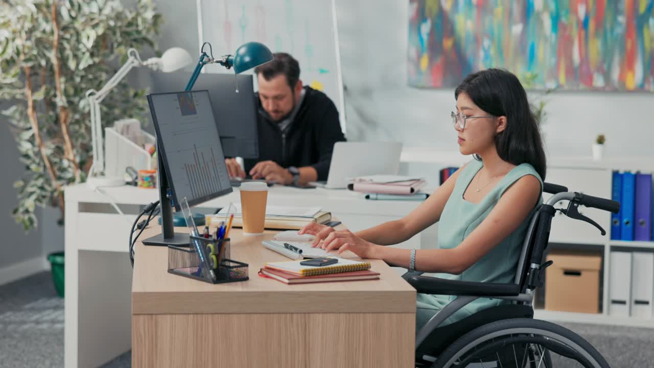 工作日在办公室，公司，公司，同事们都在电脑前工作，起草文件，一个戴着眼镜的亚洲美女坐在轮椅上，她是残疾人，事故后