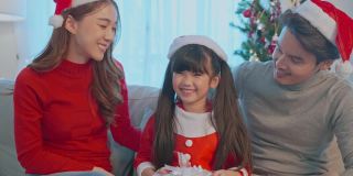 亚洲幸福家庭的肖像戴着圣诞老人的帽子，坐在客厅里。漂亮迷人的父母，穿着红衣服的爸爸妈妈和年幼的女儿看着摄像机准备在家里庆祝圣诞节