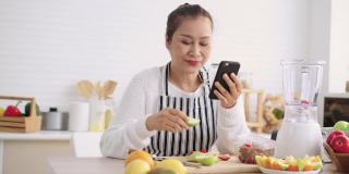 亚洲资深女性一边吃水果一边用手机感到幸福，准备用不同的口味和酸奶自制水果冰沙。健康的天然益生菌调味饮料，在厨房学习使用技术，正面视图