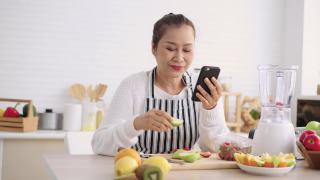 亚洲资深女性一边吃水果一边用手机感到幸福，准备用不同的口味和酸奶自制水果冰沙。健康的天然益生菌调味饮料，在厨房学习使用技术，正面视图视频素材模板下载