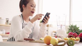 亚洲资深女性一边吃水果一边用手机感到幸福，准备用不同的口味和酸奶自制水果冰沙。健康天然益生菌调味饮料，在厨房学习使用科技视频素材模板下载