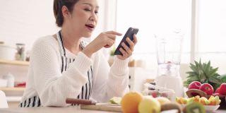 亚洲资深女性一边吃水果一边用手机感到幸福，准备用不同的口味和酸奶自制水果冰沙。健康天然益生菌调味饮料，在厨房学习使用科技