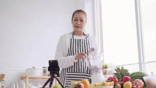 亚洲资深女性作为影响者，教如何制作自制的水果冰沙与不同的口味和酸奶。健康天然益生菌调味饮料，学会使用科技，在厨房用智能手机与观众交谈，低角度视频素材模板下载