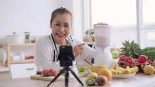亚洲资深女性作为影响者，教如何制作自制的水果冰沙与不同的口味和酸奶。健康的天然益生菌口味饮料，学习使用科技，在厨房用智能手机与观众交谈视频素材模板下载