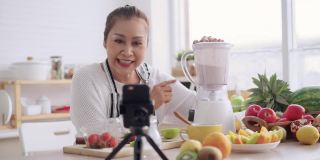 亚洲资深女性作为影响者，教如何制作自制的水果冰沙与不同的口味和酸奶。健康的天然益生菌口味饮料，学习使用科技，在厨房用智能手机与观众交谈