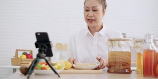 亚洲资深女性影响者教授如何制作自制的发酵生康普茶与不同的口味。健康的天然益生菌口味饮料，学习使用科技，在厨房用智能手机与观众交谈