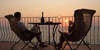 漂亮的年轻夫妇在阳台上喝着酒，欣赏着海上的日落