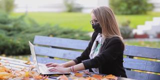 戴着新冠口罩的年轻女商人在公园里用笔记本电脑工作